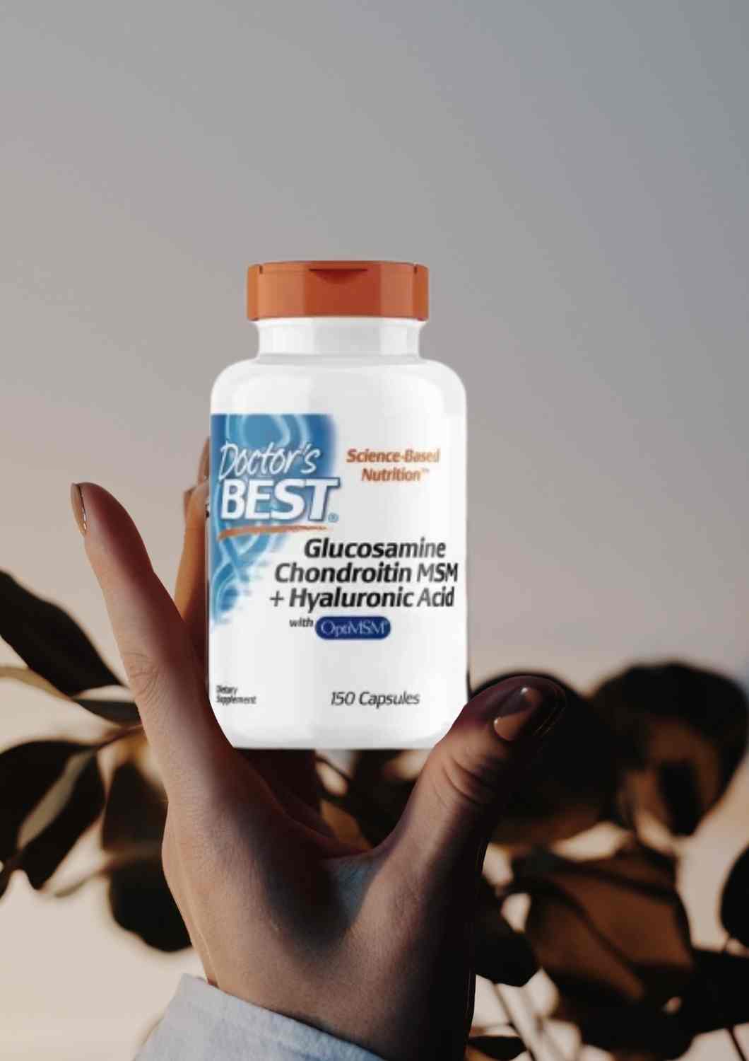 Frasco de suplemento Doctor’s Best Glucosamine Chondroitin MSM + Hyaluronic Acid sostenido por una mano, con fondo difuminado de hojas al atardecer.